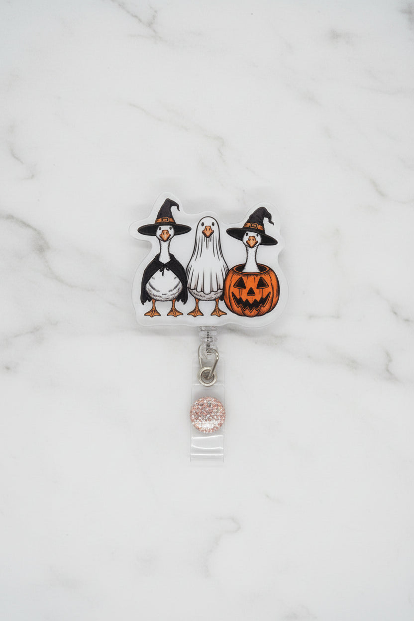 A Halloween-themed badge reel featuring three cartoon geese wearing witch hats, one ghost, and one jack-o'-lantern, attached to a white card and clear plastic sleeve on a glittery gold background.