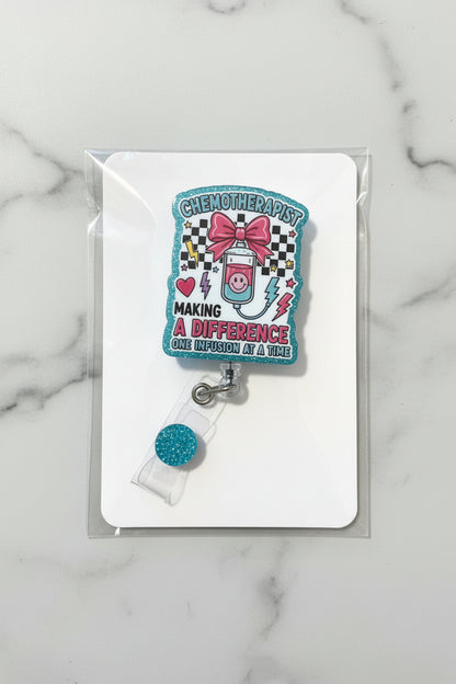 A chemotherapy-themed acrylic keychain or badge holder attached to a clear plastic clip with a blue glitter round charm, displayed on a white backing card over a gold glitter background.