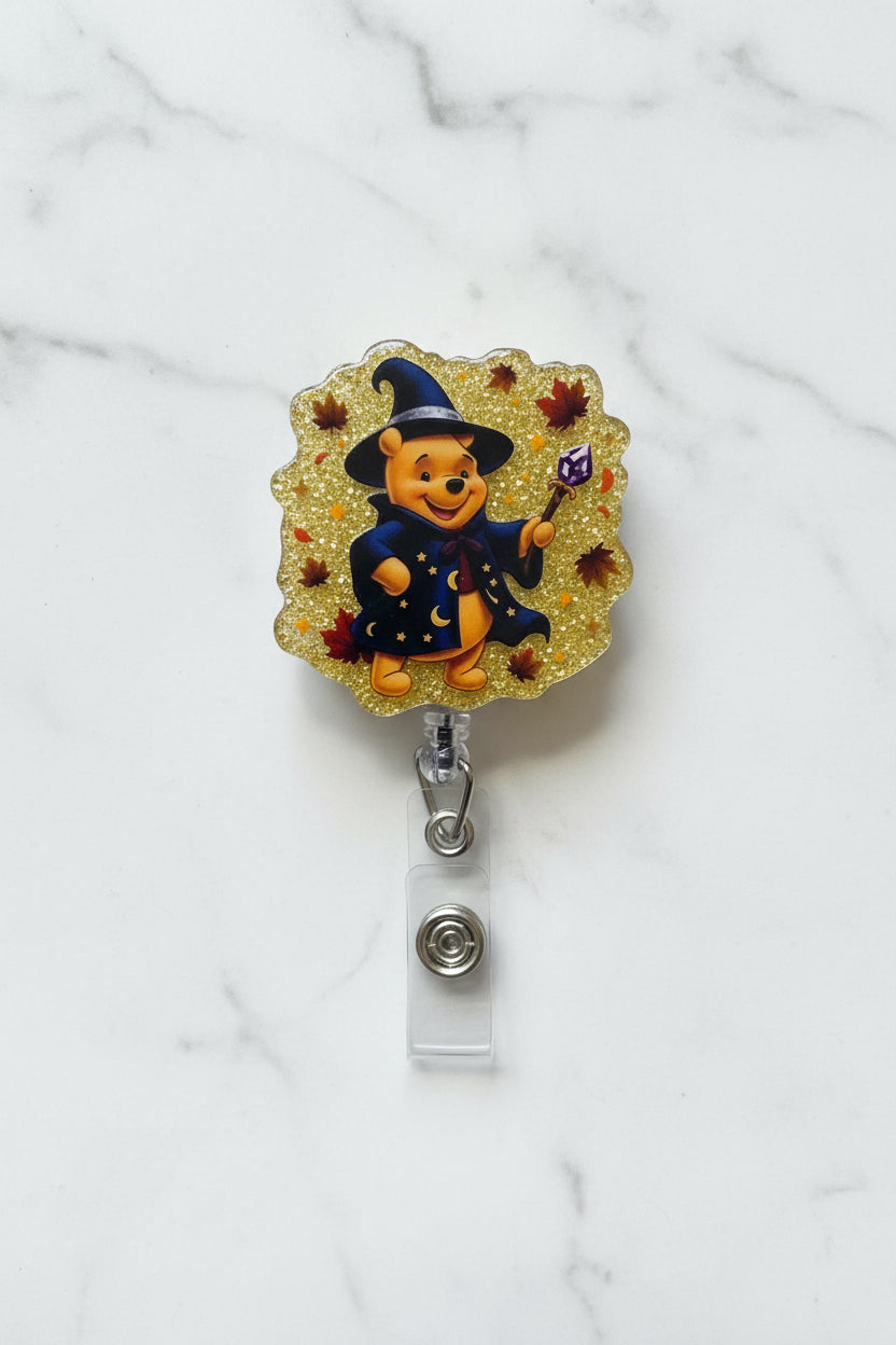 A Halloween-themed Winnie the Pooh badge reel featuring Pooh wearing a witch hat and coat on a glittery gold background, mounted on a white card inside a clear plastic sleeve.
