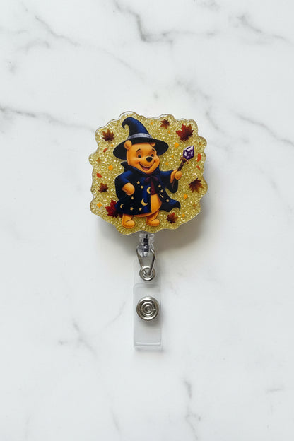 A Halloween-themed Winnie the Pooh badge reel featuring Pooh wearing a witch hat and coat on a glittery gold background, mounted on a white card inside a clear plastic sleeve.